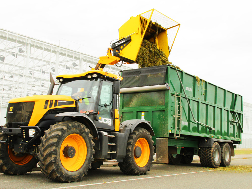JCB tractor loading greenhouse plant waste into a green trailer beside a glasshouse